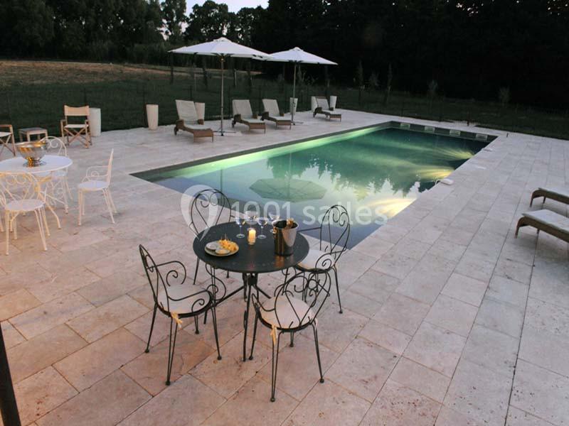 Terrasse en pierre avec piscine éclairée au crépuscule, entourée de chaises longues, tables et parasols.