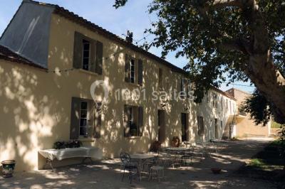 Façade d'une maison provençale avec terrasse ombragée, tables en fer forgé et grand arbre au premier plan.