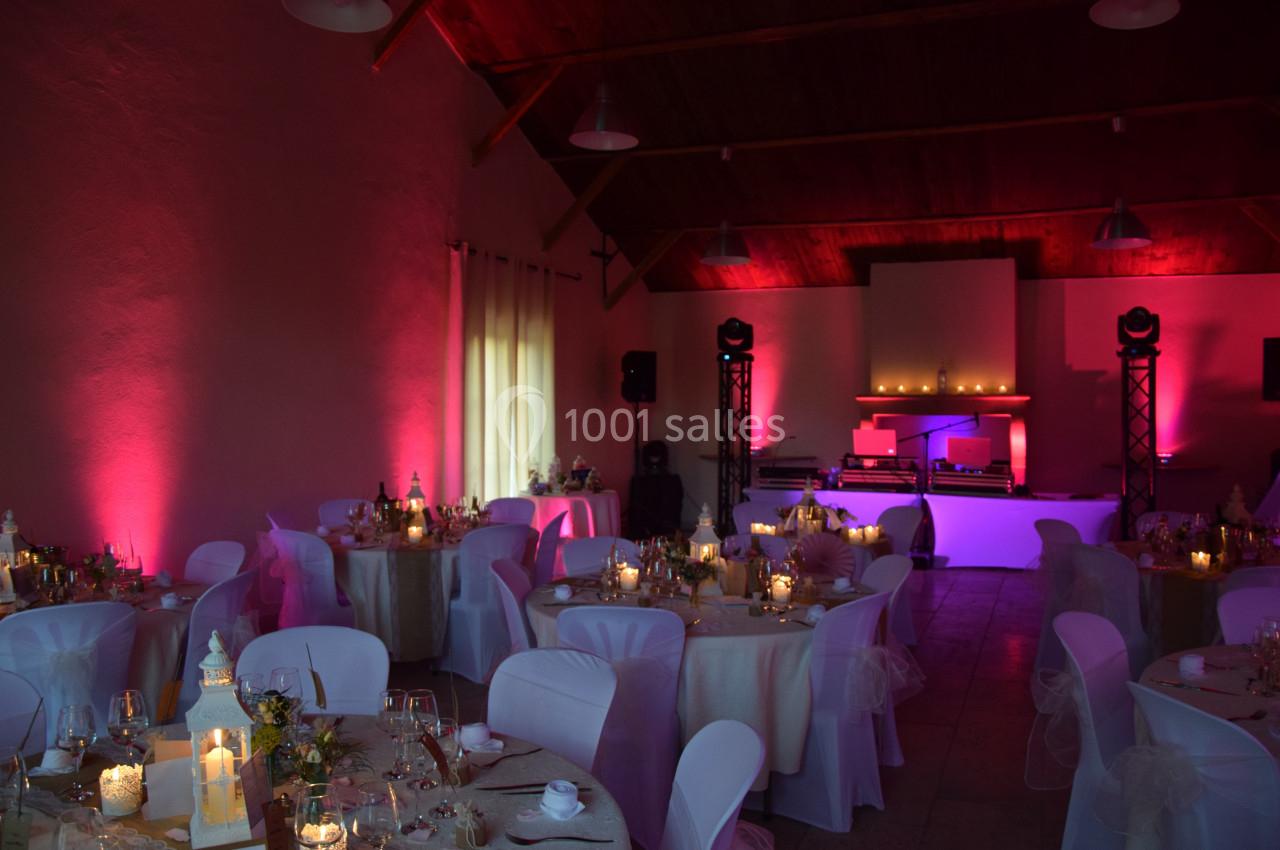 Salle de réception décorée avec des tables rondes, nappes blanches, bougies et éclairage d'ambiance rose et violet.