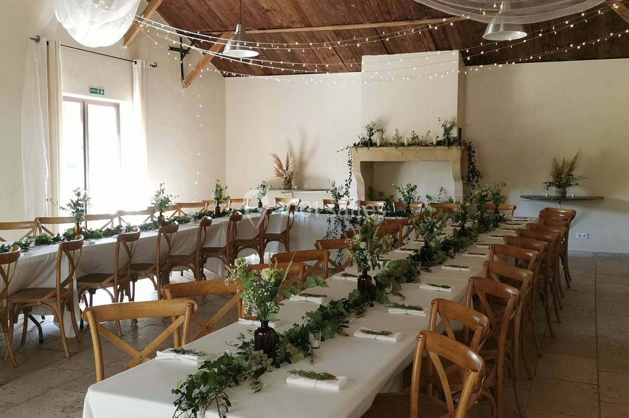 Salle de réception décorée avec des tables alignées, nappes blanches, guirlandes végétales et voilages suspendus.