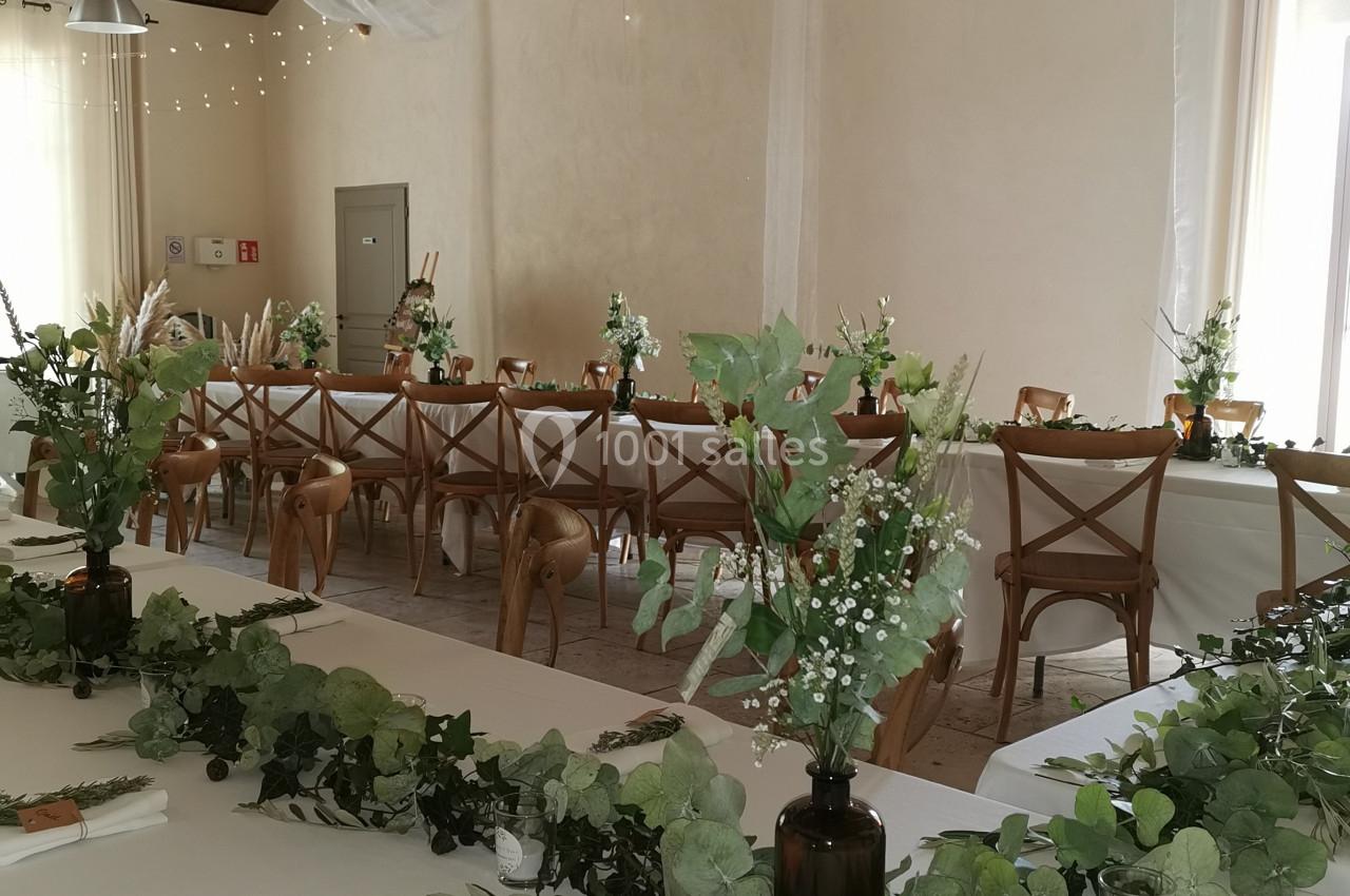 Salle décorée pour un événement avec tables blanches, chaises en bois, guirlandes lumineuses et compositions végétales.