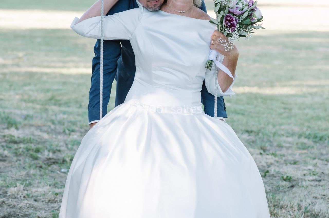 Un couple de mariés souriants, la mariée tenant un bouquet, assis sur une balançoire dans un parc verdoyant.