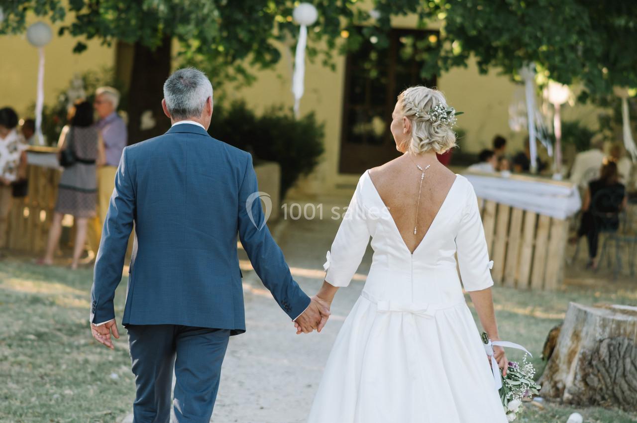 Un couple en tenue de mariage marche main dans la main sur un chemin bordé d'herbe, devant des invités en arrière-plan.