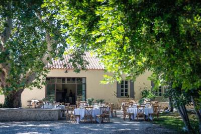Façade d'une maison provençale avec terrasse ombragée, tables en fer forgé et grand arbre au premier plan.