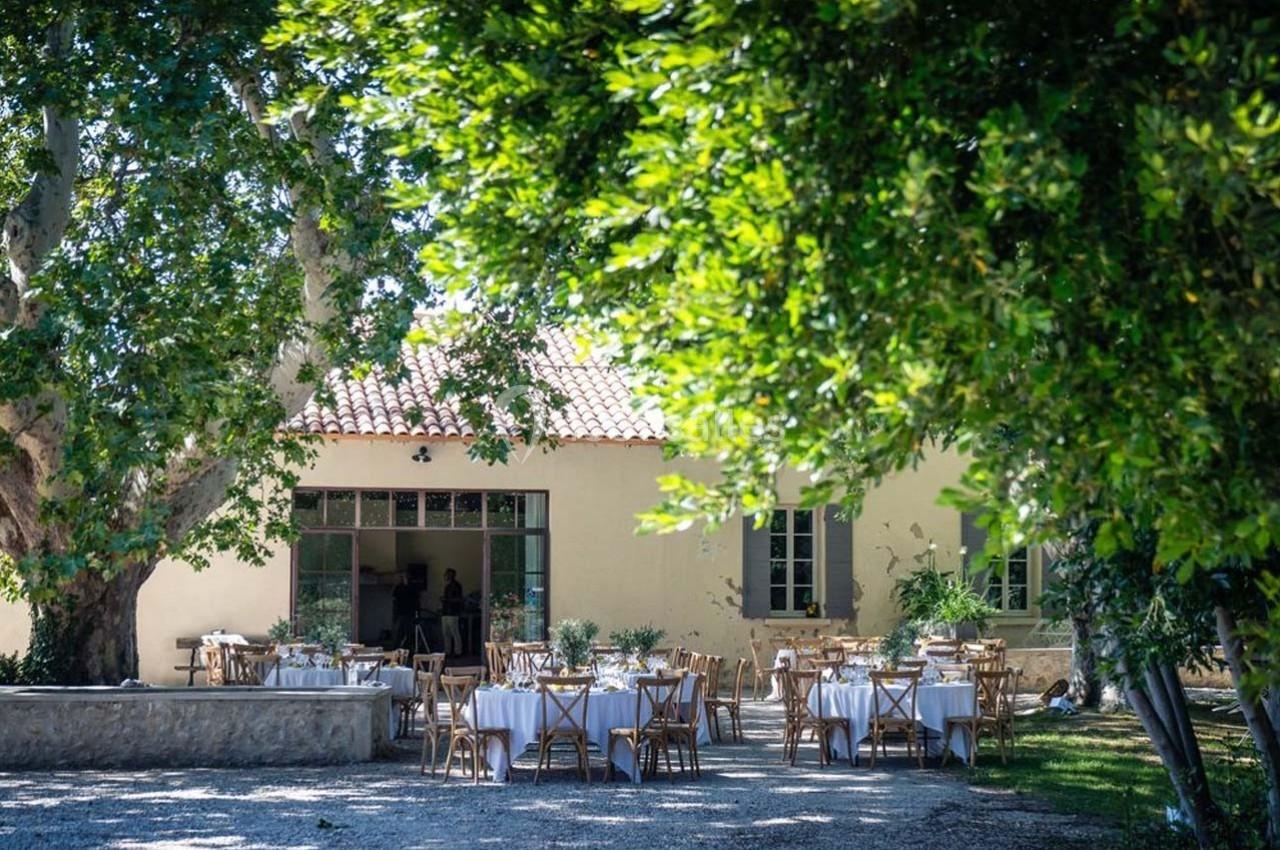 Tables dressées en extérieur sous des arbres, devant une maison aux murs clairs avec des fenêtres à volets verts.