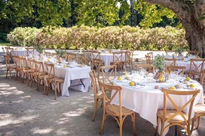 Façade d'une maison provençale avec terrasse ombragée, tables en fer forgé et grand arbre au premier plan.