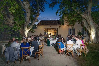 Façade d'une maison provençale avec terrasse ombragée, tables en fer forgé et grand arbre au premier plan.