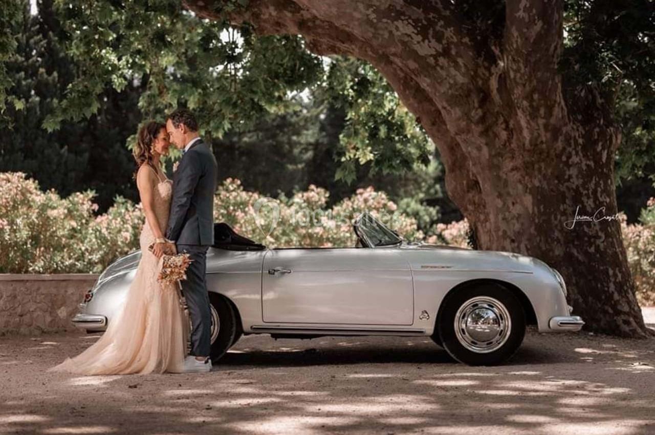Un couple habillé élégamment se tient près d'une voiture ancienne sous un grand arbre, dans un cadre naturel.