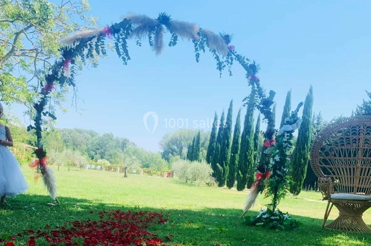 Arche décorée de fleurs et de feuillages sur une pelouse, avec un fauteuil en osier et des pétales rouges au sol.