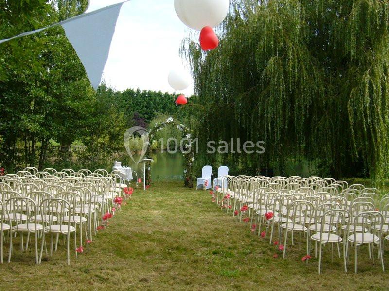 Chaises alignées en extérieur pour une cérémonie, avec arche décorée et ballons, près d'un étang entouré de verdure.