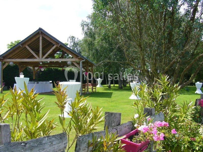 Abri en bois dans un jardin verdoyant avec tables hautes blanches et fleurs en premier plan.