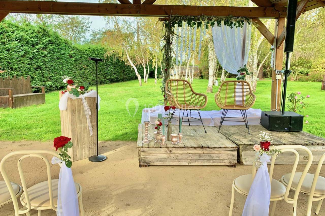Espace extérieur décoré pour une cérémonie, avec chaises blanches, podium fleuri et estrade sous une pergola en bois.