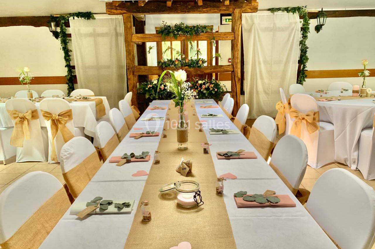 Salle décorée pour un événement, avec longues tables ornées de nappes blanches, chemins de table dorés et chaises habillées.