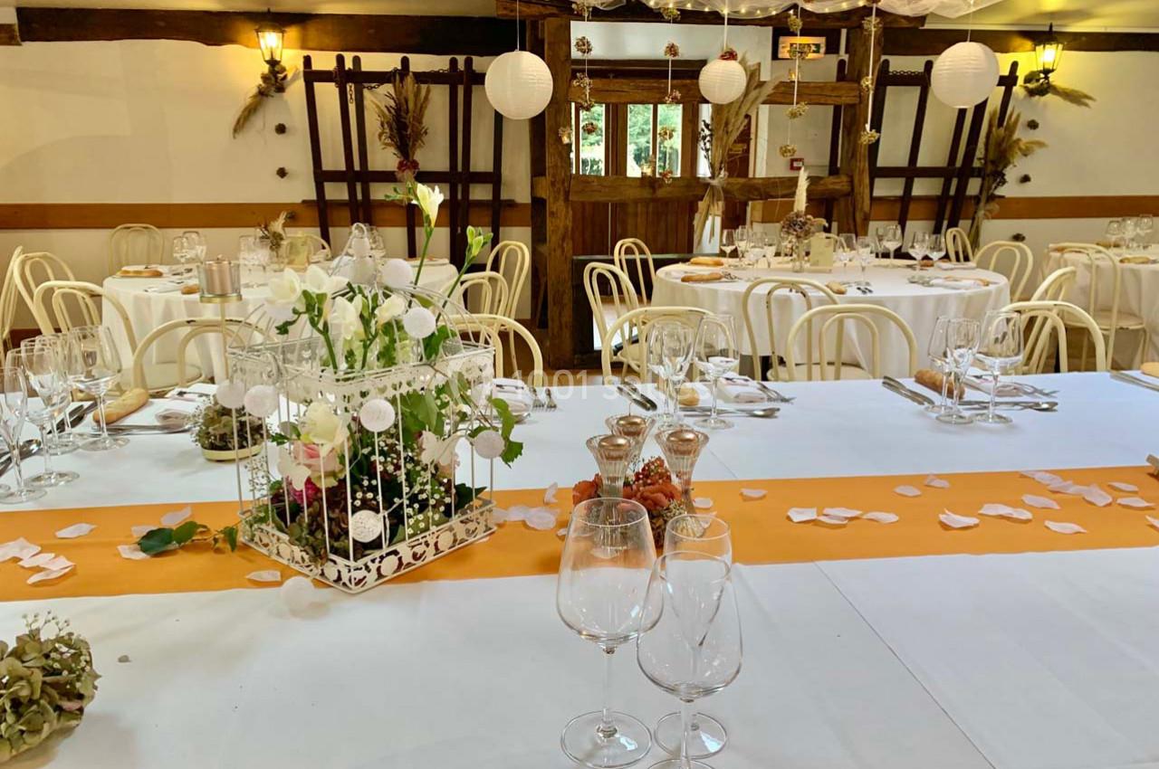Salle décorée pour un événement avec tables dressées, nappes blanches, chemins de table orange et centre de table floral.