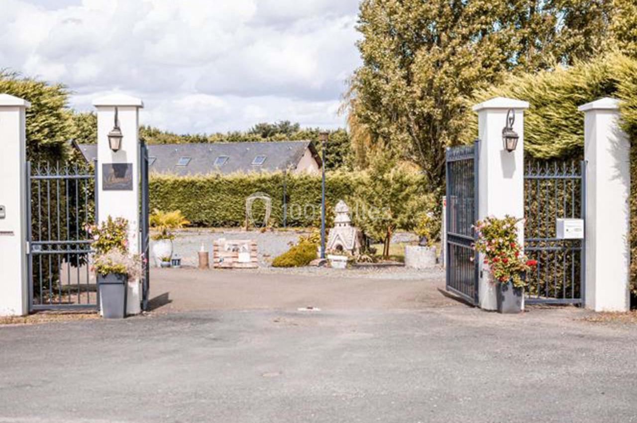 Entrée d'une propriété avec portail ouvert, colonnes blanches, pots de fleurs et jardin arboré en arrière-plan.