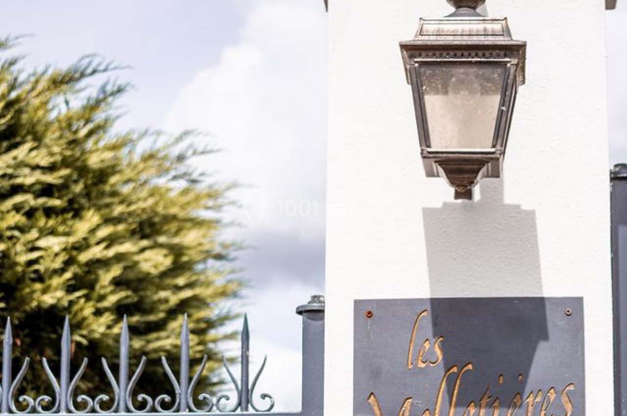 Lampe murale ancienne fixée sur un pilier blanc, avec une plaque indiquant ’Les Melletières’ et une grille en fer forgé.