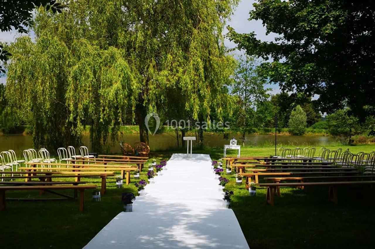 Allée centrale blanche entourée de bancs et chaises en bois, installée en extérieur sous des arbres près d'un plan d'eau.