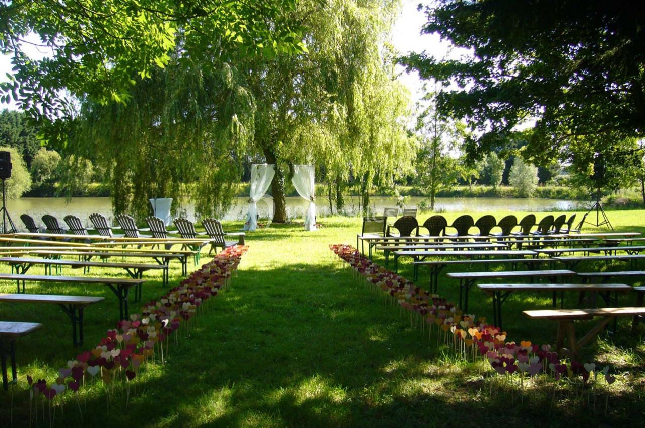 Allée bordée de fleurs menant à un espace de cérémonie en plein air, avec chaises alignées et vue sur un étang.