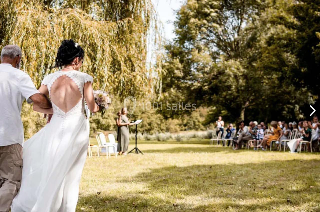 Une mariée en robe blanche marche dans un parc verdoyant, accompagnée d'un homme, vers une cérémonie en plein air.