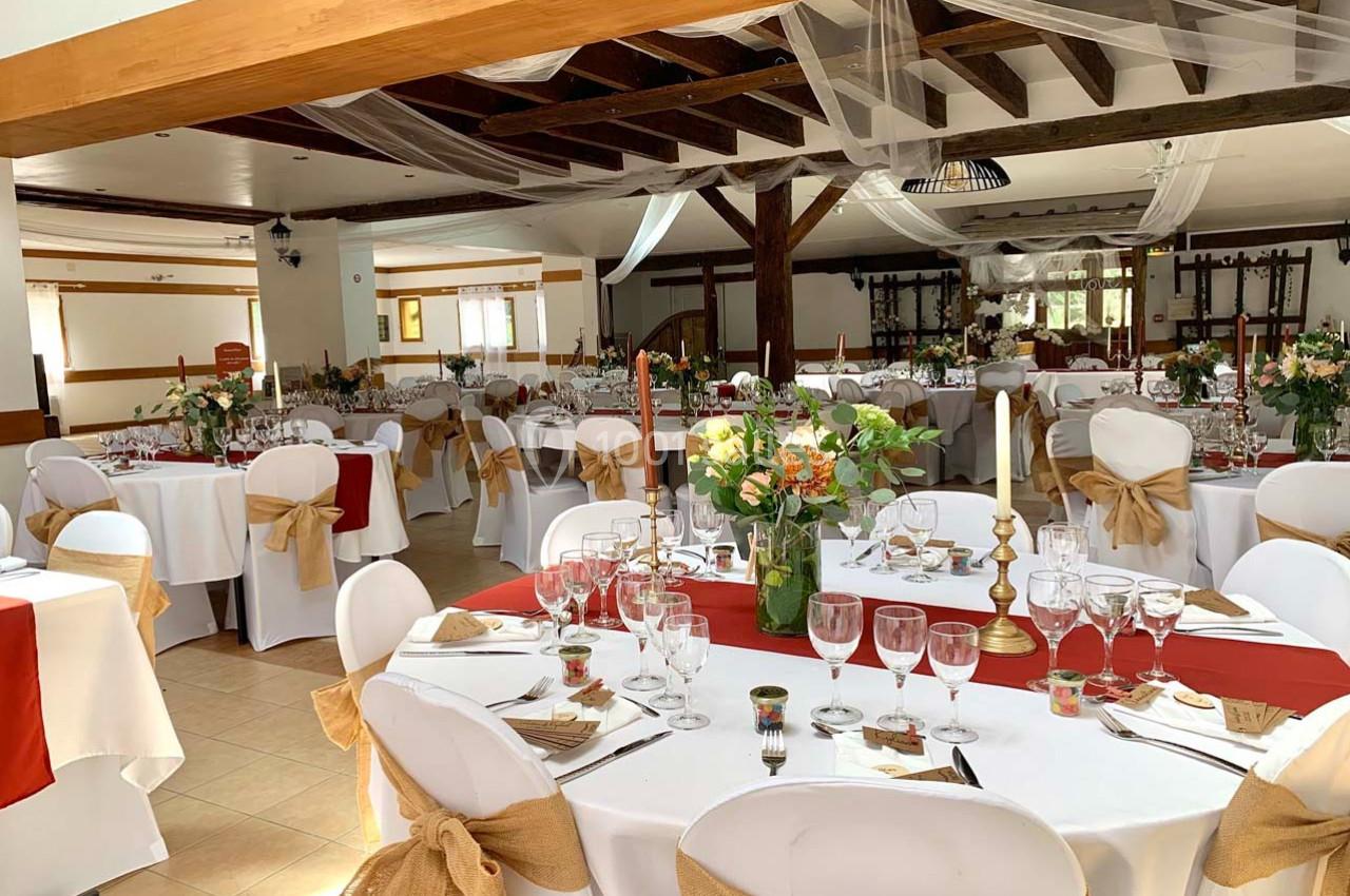 Salle de réception décorée pour un événement, avec tables dressées, nappes blanches, chemins de table rouges et chaises…