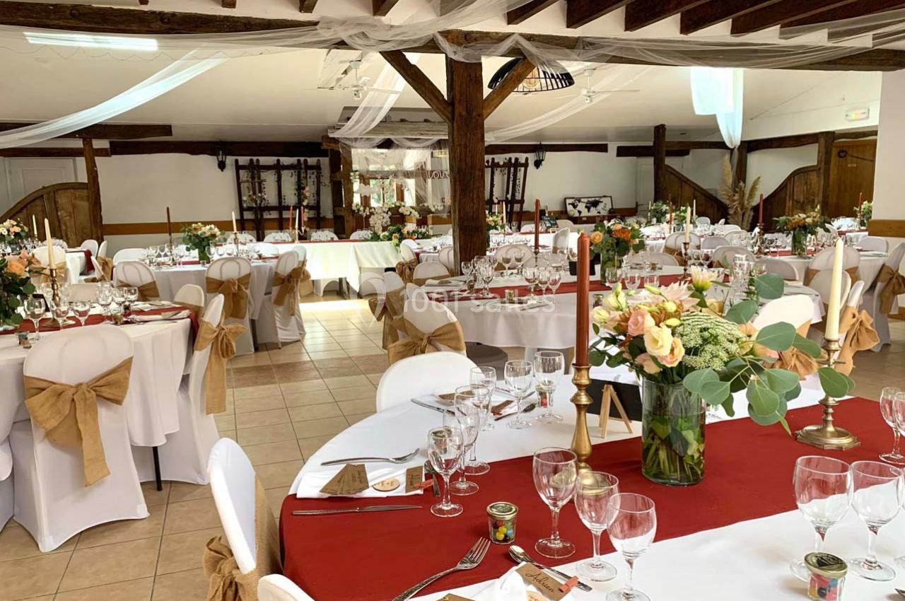 Salle de réception décorée avec des tables rondes, nappes blanches, chemins de table rouges et chaises ornées de nœuds…