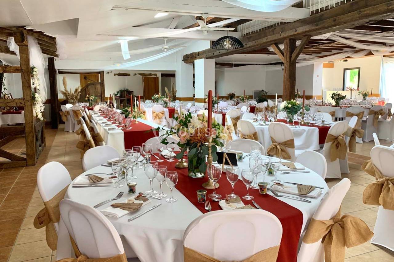 Salle de réception décorée pour un événement, avec tables rondes dressées, nappes blanches et chemins de table rouges.