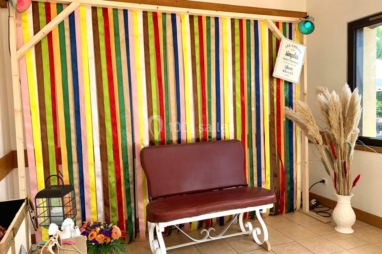 Banc rouge devant un mur décoré de bandes colorées, avec des fleurs, une lanterne et des herbes séchées à proximité.