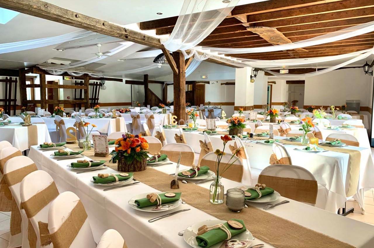 Salle de réception décorée pour un événement, avec tables dressées, nappes blanches, chemins de table en jute et fleurs…