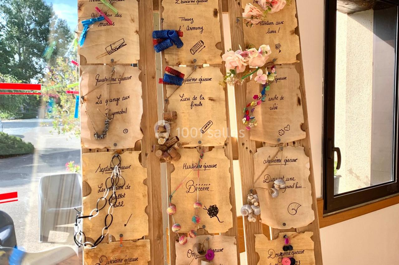 Panneau en bois décoré de parchemins manuscrits, rubans, fleurs et petits objets, exposé près d'une fenêtre.