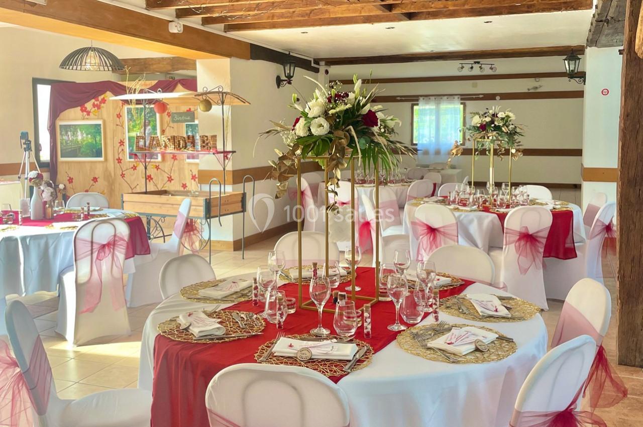 Salle de réception décorée avec des tables rondes, nappes blanches, chemins de table rouges et centres de table fleuris.