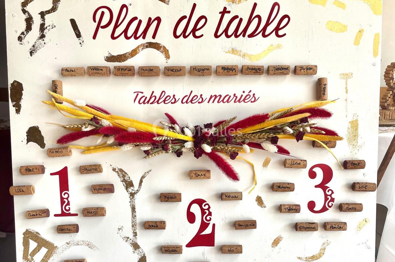 Plan de table décoratif avec des noms inscrits sur des bouchons de liège, orné de fleurs séchées rouges et jaunes.