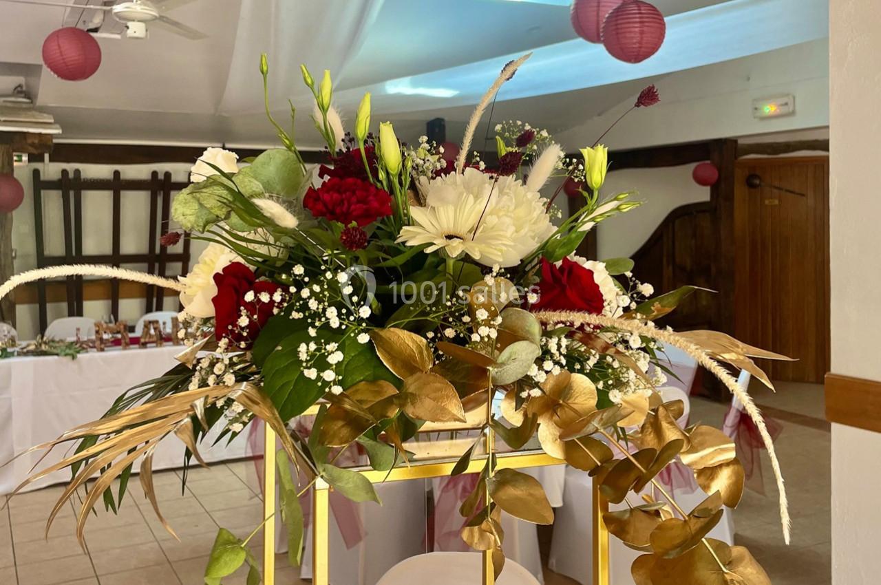 Arrangement floral avec roses rouges, fleurs blanches, gypsophile et feuilles dorées sur un support doré dans une salle…