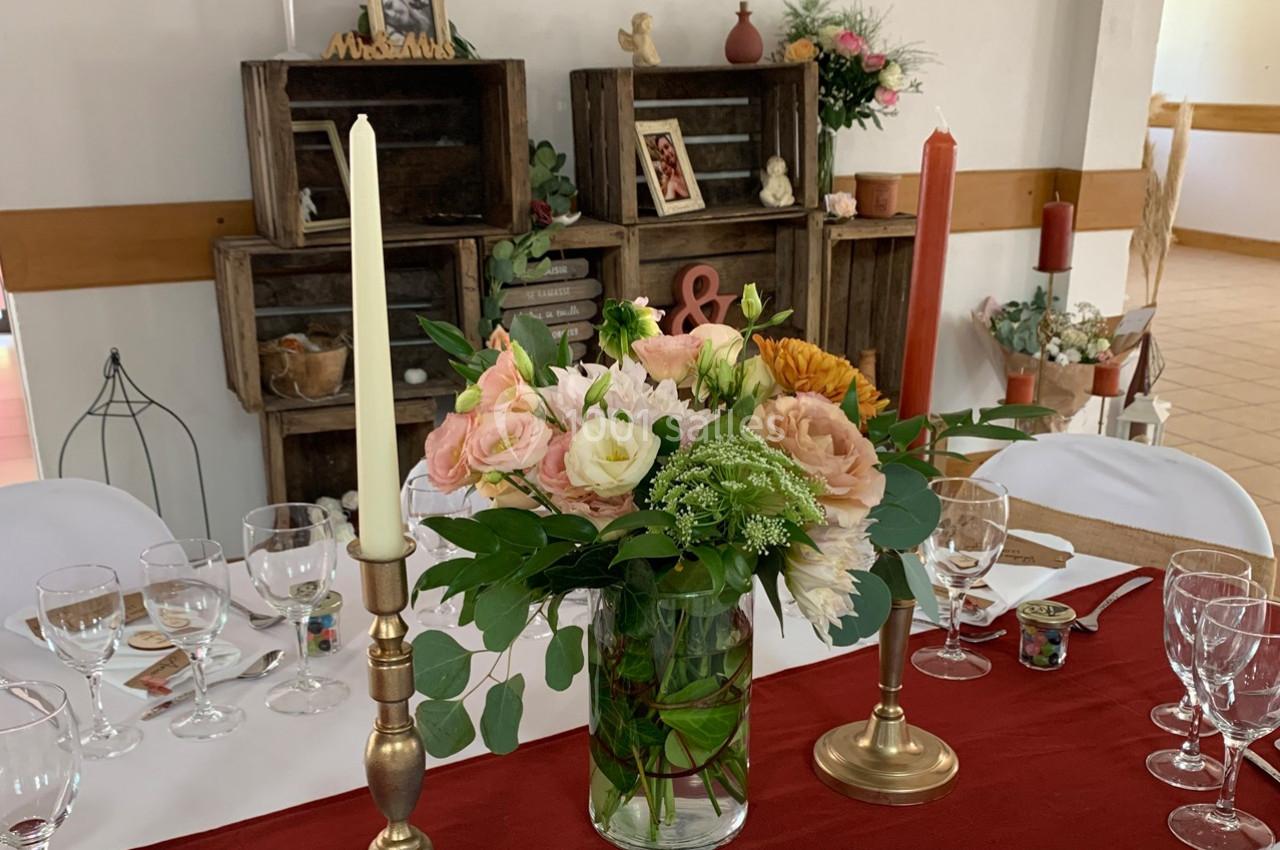 Centre de table avec bouquet de fleurs pastel, chandeliers dorés et décor rustique en arrière-plan.