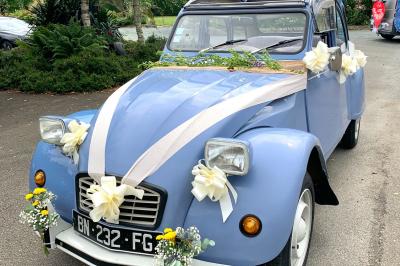 Voiture ancienne bleue décorée de rubans blancs et de fleurs pour une cérémonie, stationnée en extérieur.