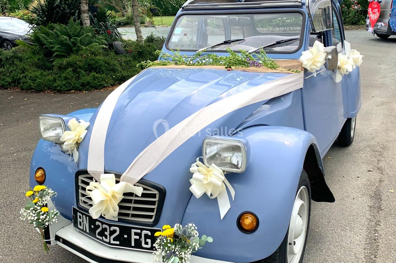 Voiture ancienne bleue décorée de rubans blancs et de fleurs pour une cérémonie, stationnée en extérieur.