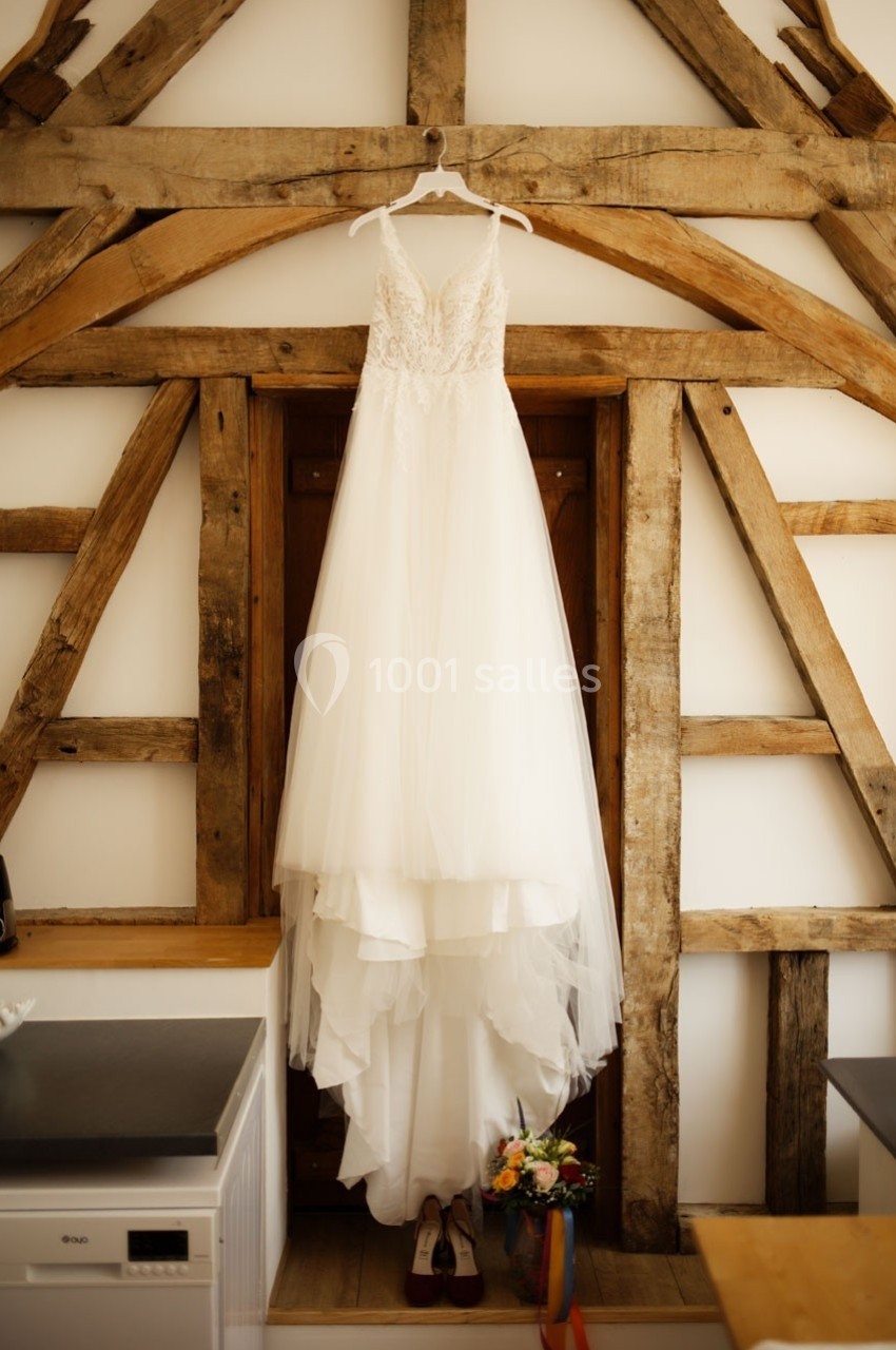 Robe de mariée blanche suspendue devant une porte en bois encadrée de poutres apparentes, avec des fleurs et des chaussures…