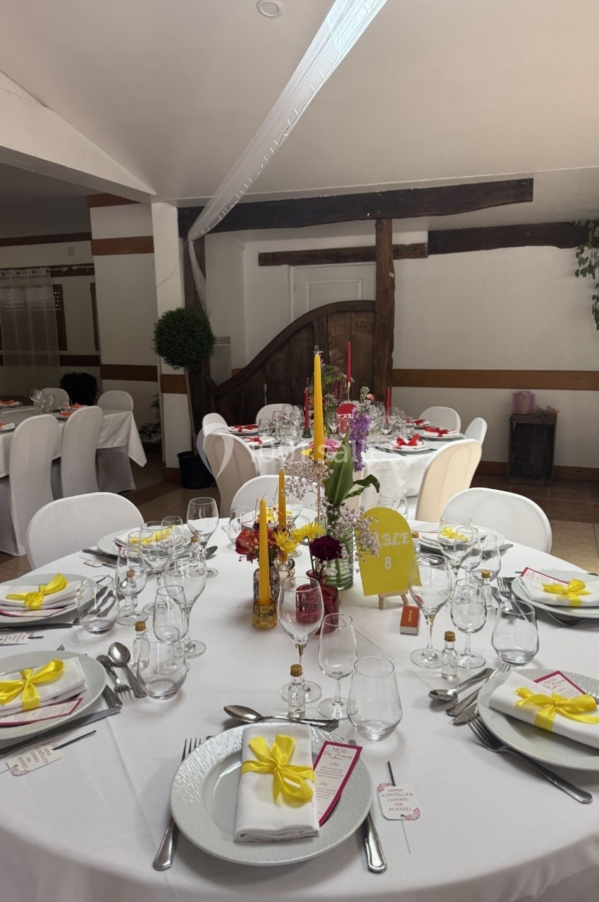Salle décorée pour un événement avec tables rondes dressées, nappes blanches, serviettes nouées de rubans jaunes et centre…