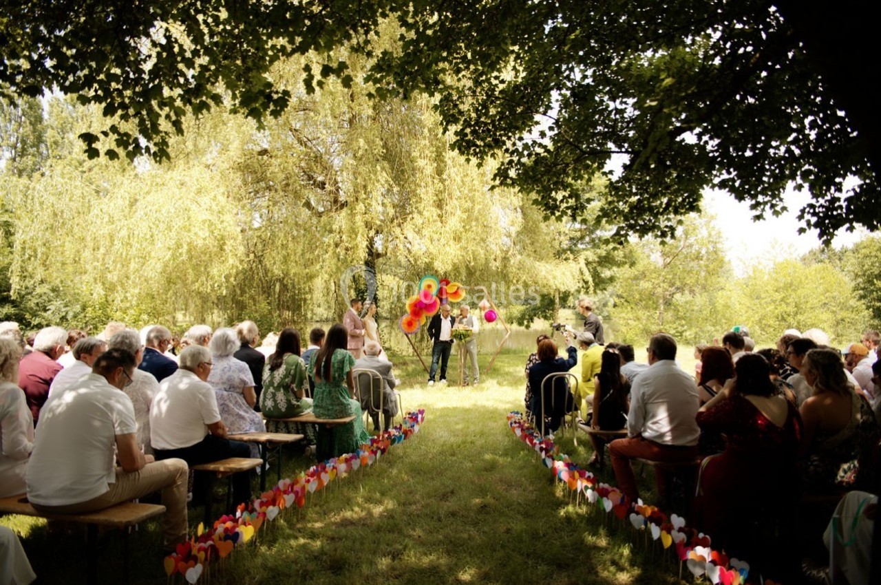 Cérémonie en plein air avec des invités assis sur des bancs, allée décorée de cœurs et ballons colorés au centre.