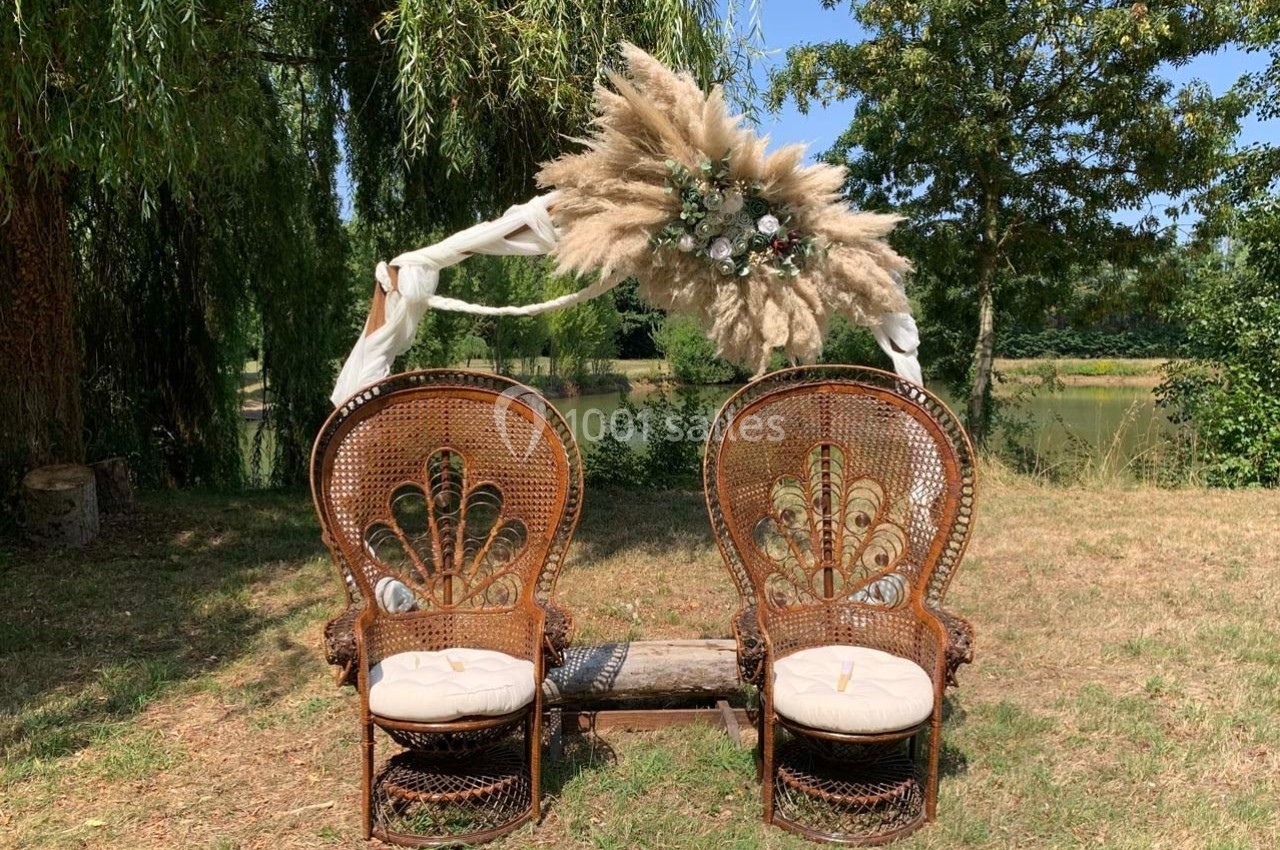 Deux fauteuils en rotin ornés de coussins, disposés sous une arche décorée de fleurs et de pampas, en extérieur.