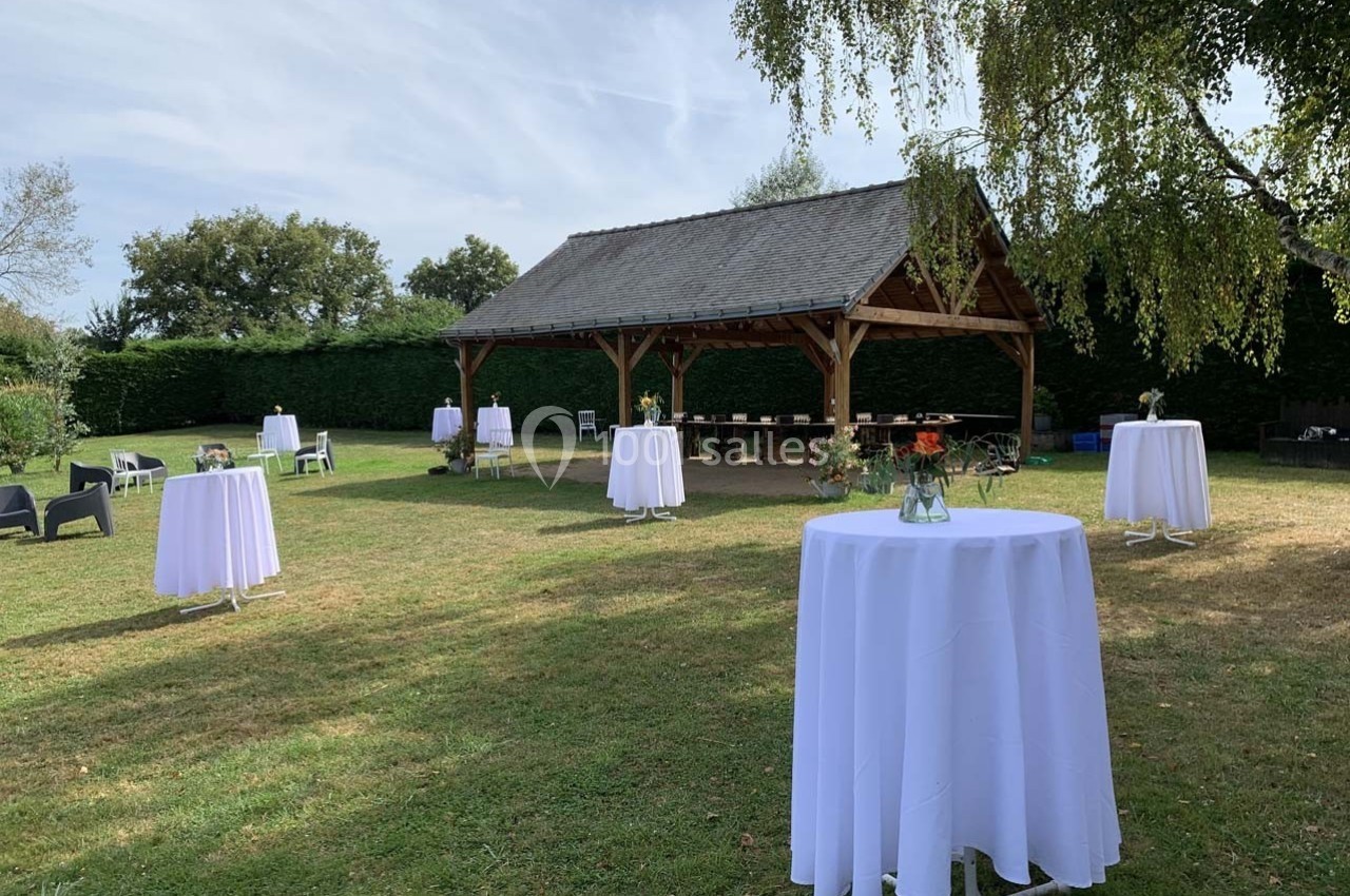 Tables hautes dressées avec nappes blanches dans un jardin, près d'un abri en bois, sous un ciel dégagé.