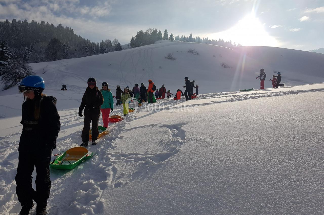 Des enfants et adultes alignés avec des luges sur une pente enneigée, sous un ciel ensoleillé.