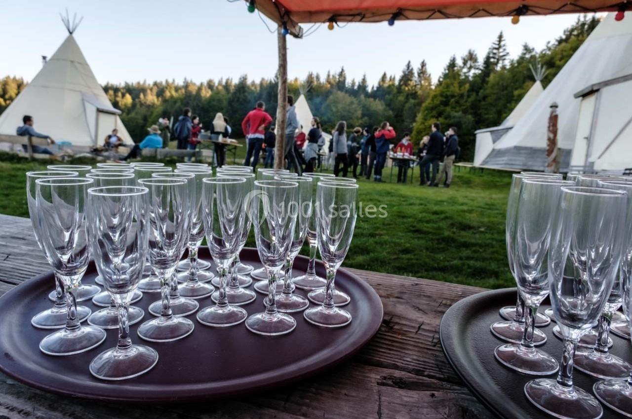 Plateaux de verres à pied posés sur une table en bois, avec des tipis et des personnes en arrière-plan dans un cadre naturel.
