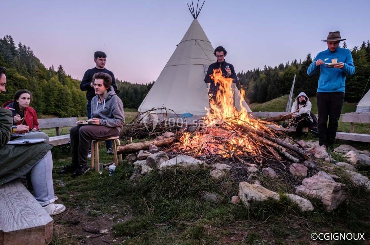 Des personnes dînent autour d'un feu de camp, avec des tipis en arrière-plan dans un paysage naturel.
