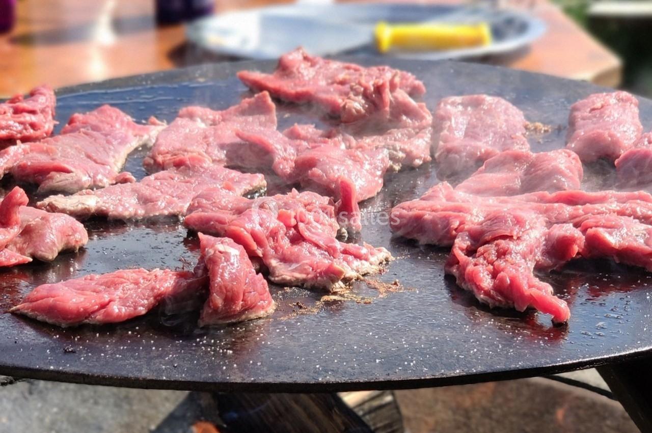 Viande rouge en train de griller sur une plancha en extérieur, avec des ustensiles visibles en arrière-plan.