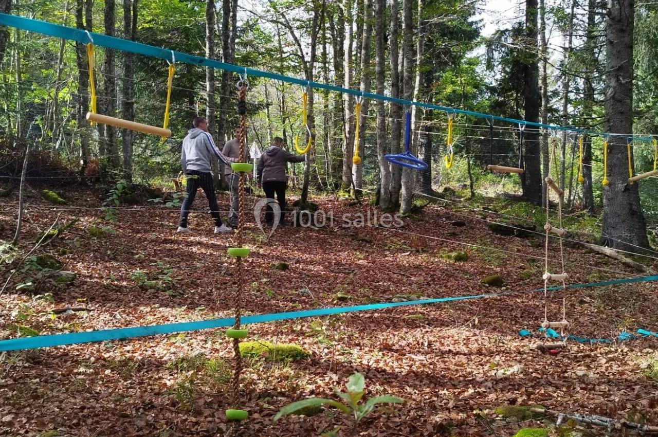 Deux personnes traversent un parcours d'accrobranche en forêt, entourées d'arbres et de feuilles au sol.