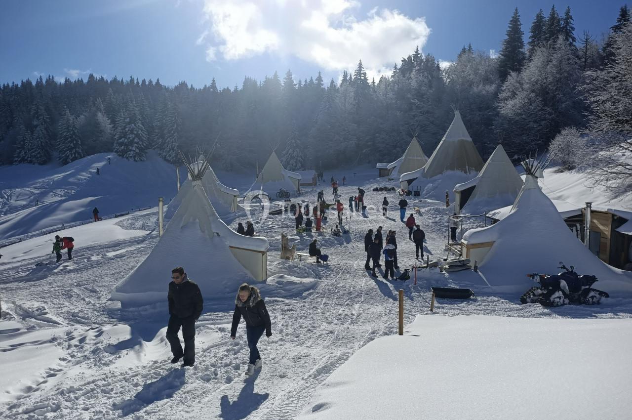 Personnes marchant et se rassemblant autour de tipis enneigés dans un paysage de montagne sous un ciel ensoleillé.