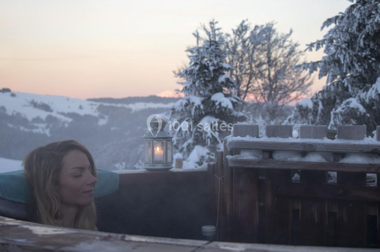 Femme se relaxant dans un bain chaud en extérieur, entourée de neige et de sapins sous un ciel au coucher du soleil.