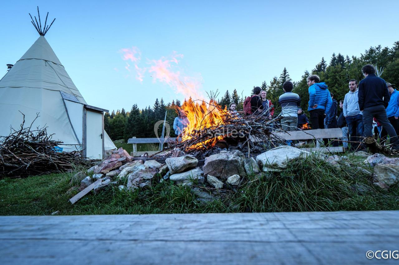 Un groupe de personnes rassemblé autour d'un feu de camp près d'un tipi dans un paysage forestier au crépuscule.