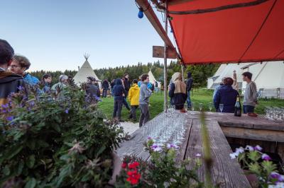 Groupe de personnes rassemblées en extérieur près de tentes et d'une table avec des verres, dans un cadre naturel.