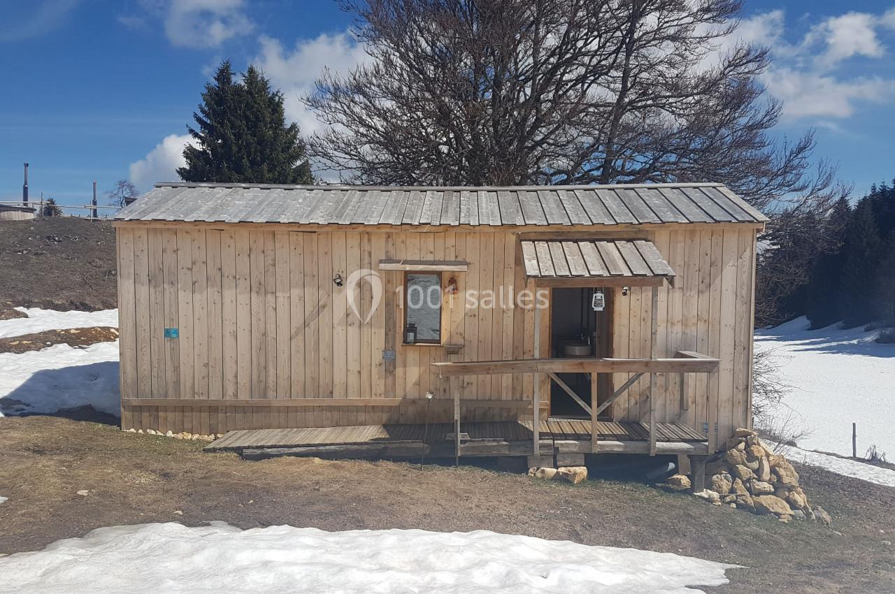 Cabane en bois sur un terrain partiellement enneigé, entourée d'arbres et sous un ciel bleu.